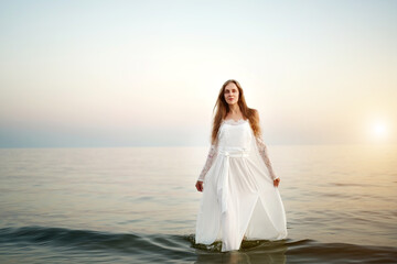 Fototapeta na wymiar Perfect beautiful girl emerges from a calm sea at sunset. Summer nature, serenity. Very cute, clear, elegant clothes: white lace dress, transparent sleeves, a long skirt that flutters in the wind.