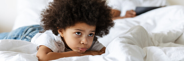 Black boy frowning while his father using tablet computer in bed