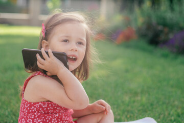 Portrait of little 3-4 girl in red clothes sitting on blanket on green grass and speaking on cell phone. Children using gadgets