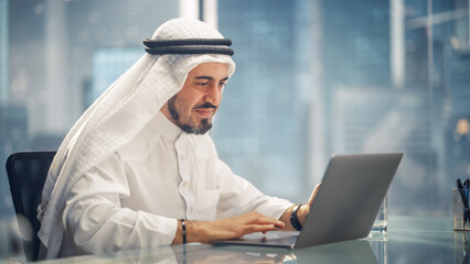 Arab Businessman in White Traditional Outfit Sitting in Office and Working on Laptop Computer....