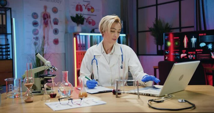 Medicine concept where beautiful concentrated busy skilled blond woman doctor working with computer information and paper reports during overtime work in evening lab