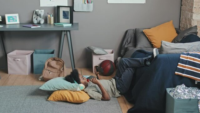 Long side view of ten-year-old Black boy wearing over-ear headphones, lying on floor with legs up on couch in his room at daytime, having lollipop and using smartphone