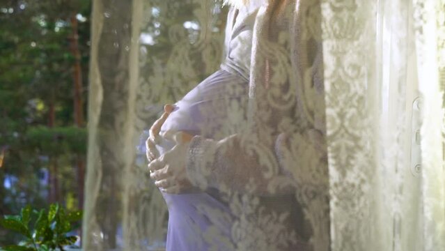 Pregnant Woman standing on the balcony in front of an open window and stroking her big belly with love, looking at the pine forest outside the window. Motherhood, future life and ecology concept