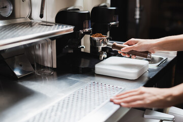 Cropped view of barista holding portafilter near grinder and coffee machine in cafe.