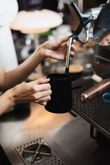 Cropped view of barista holding milk jug near steam wand of coffee machine in cafe.