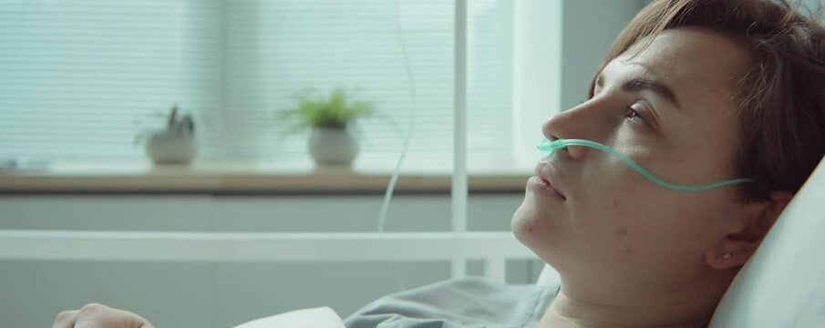 Young ill woman with nasal cannula sleeping on bed in hospital ward, opening her eyes and looking at side