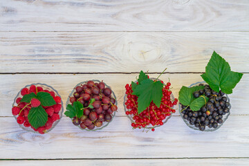 Raspberries, red currants, gooseberries, black currants in glass plates on a rustic white wooden table
