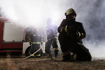 Team of professional firefighters holding water hose in front of a firetruck with smoke in the air. Half silhouette.