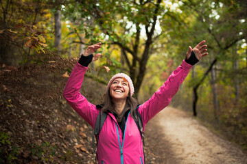 Hiking girl in pink on a trail in the forest. Hands up enjoying the falling leaves in nature in fall season.