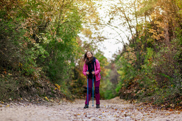Naklejka na ściany i meble Hiker girl standing on a wide trail in the mountains. Backpacker with pink jacket in a forest. Healthy fitness lifestyle outdoors.