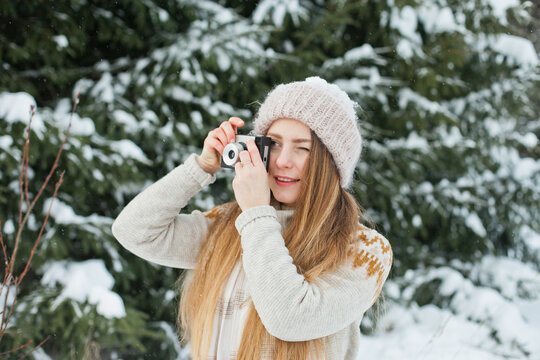 Beautiful millennial young woman taking pictures with retro vintage film camera. Hobby and leisure activity in winter forest outside. Amateur photographer taking photos with snow.