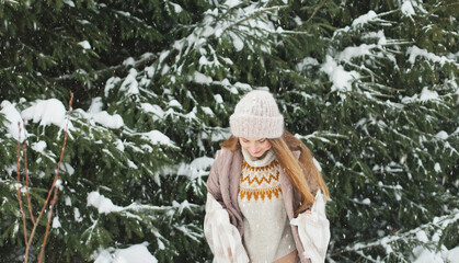 Happy woman in winter forest during snowfall, enjoying winter. Millennial young woman with long hair in beige warm outfit walking in the snowy park in nature near fir trees, being careless and free..
