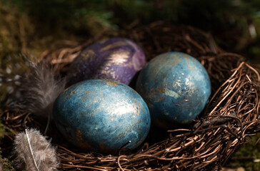 Happy Easter 2022. A postcard with a copy of the place for the text. Purple, blue and golden eggs in a nest on a natural forest background. Top view.