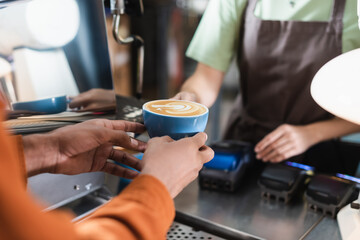 Cropped view of african american man holding coffee near blurred barista in cafe.
