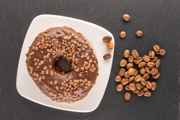 One sweet chocolate donut on a white saucer with coffee beans, on a slate rock , macro, top view.