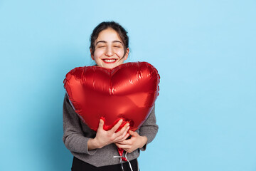 Nice young smiling woman with natural make up standing with heart balloon isolated on blue background. Valentines Day concept