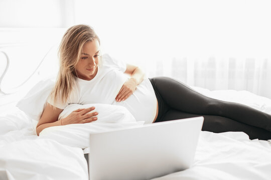 Young pregnant woman watching film on laptop lying on bed