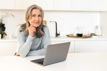 Cheerful attractive mature woman looks sits in the kitchen with laptop, happy senior female freelancer chatting online, enjoying remote work