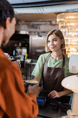 Smiling barista holding coffee near blurred african american customer in cafe.