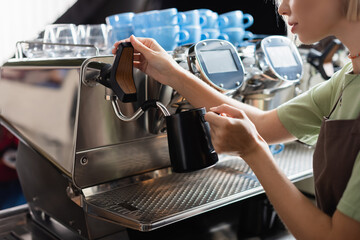 Cropped view of barista holding milk jug near coffee machine with steam wand in cafe.
