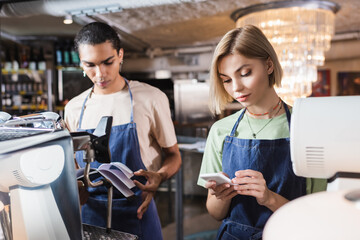 Young barista using smartphone near african american colleague with notebook in cafe.