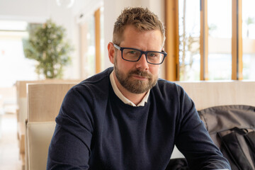 Portrait of a bearded man in glasses looking at camera sitting in café or coffeeshop.
