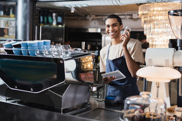 Smiling african american barista holding devices near coffee machine in cafe.