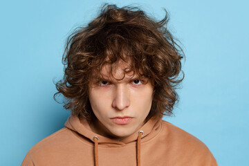 Close-up angry young man, student in brown hoodie gazes isolated on blue background. Concept of...