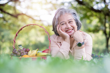 Happy Asian old senior woman and wear a health watch and lying on the picnic mat in park and basket...
