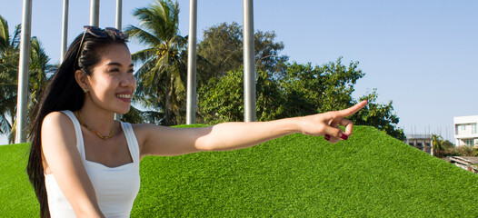 Young asian woman sits on the lawn in the park and waving her hand in greeting