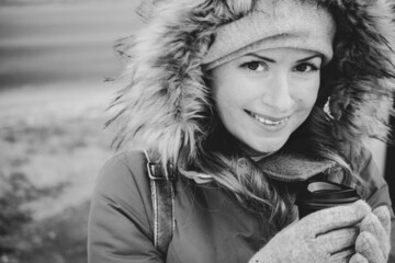 Smiling girl with coffee in warm jacket with fur hood, black and white. Pretty woman in hooded...