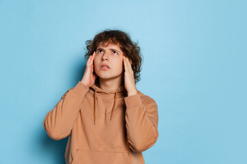 Portrait of young sad man, student in brown hoodie posing isolated on blue background. Concept of emotions, study, education, fashion