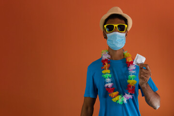 Black man in carnival costume and pandemic mask holding  a condom isolated on orange background.