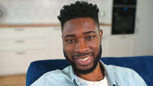 Close-up portrait of carefree African-American guy in casual wear sitting on the sofa at home and looking at the camera, feels happy, cheerful multiracial man smiling toothy