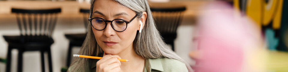Mature grey woman in earphone working with laptop and papers