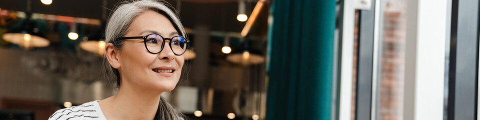 Mature woman smiling and using cellphone while sitting in cafe indoors