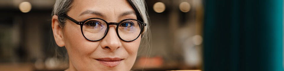 Mature grey woman wearing eyeglasses looking at camera while sitting in cafe