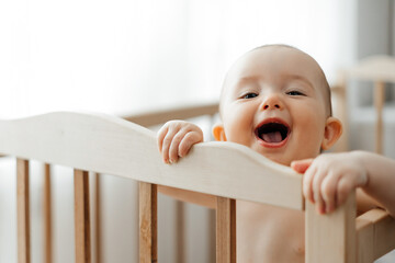 portrait of a 5 month old baby girl standing in a crib in a bright nursery after sleep and looking...