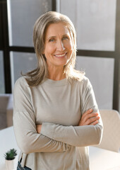 Vertical portrait of senior mature gray-haired female professional, leader, coach, elder businesswoman standing with her arms crossed folded in the office and confidently looking at the camera