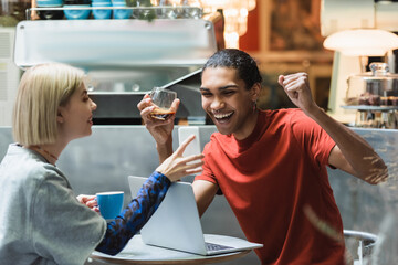 Excited african american freelancer holding coffee near girlfriend and devices in cafe.