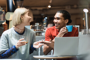 Smiling interracial freelancers talking and holding coffee near devices in cafe.
