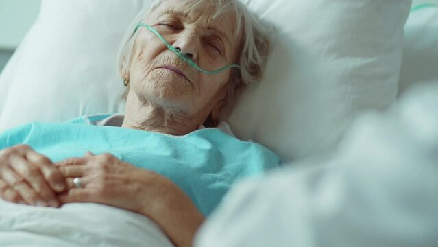 Over the shoulder shot of unconscious elderly woman with nasal cannula lying with eyes closed on hospital bed while unrecognizable doctor in gloves holding her hand