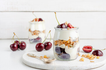 Jars of granola with yogurt and sweet cherries on white wooden board. Healthy breakfast, copy space.