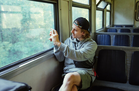 Positive young man in casual clothes rides in an old train and takes photos of beautiful views from the window.