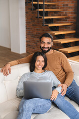 Young Pakistani couple using laptop while sitting on the sofa at home. Spouses watching movie on laptop. Happy man and woman embracing while looking at the camera