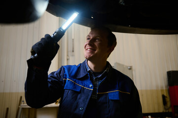 Mechanic, technician, auto engineer checks the underbody of a lifted car with a lamp in the garage of a repair shop. Car warranty repair and maintenance, car service concept