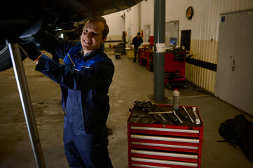 Handsome Caucasian young man, car engineer, technician, auto mechanic repairing a lifted modern automobile on a hoist in the repair shop garage. Automobile maintenance and auto service concept