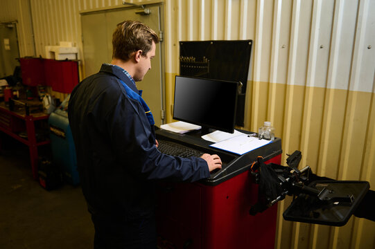 Rear view of a garage technician making check list for repairing car, typing on keyboard and using computer monitor while servicing the automobile during warranty maintenance in the auto service