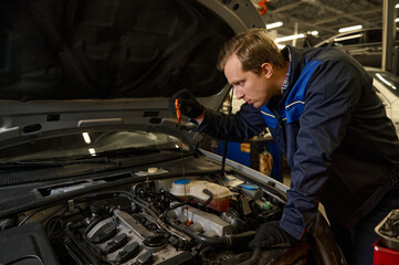 Fototapeta na wymiar Concentrated Caucasian young man, car engineer technician mechanic in professional uniform, automobile mechanic repairman testing and checking oil level in the car engine. Car service and maintenance