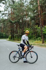 Fototapeta premium Athletic young man stands with a bicycle in the woods on the road, wears sports equipment and poses for the camera on the background of a beautiful view of nature.
